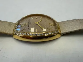 Vacheron Constantin Egerie 25540 наручные часы «Vacheron Constantin Egerie ref. 25540», корпус овальный из металла золотистого цвета со вставкой из прозрачных камней по кругу. Ремешок из материала светлого цвета с замком из металла золотистого цвета, на оборотной стороне штампованная надпись…